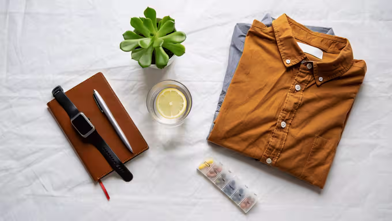 Two folded shirts, on a white bedsheet. A brown leather notebook with a silver pen. A clear glass of water with a lemon slice is positioned to the lower left. 