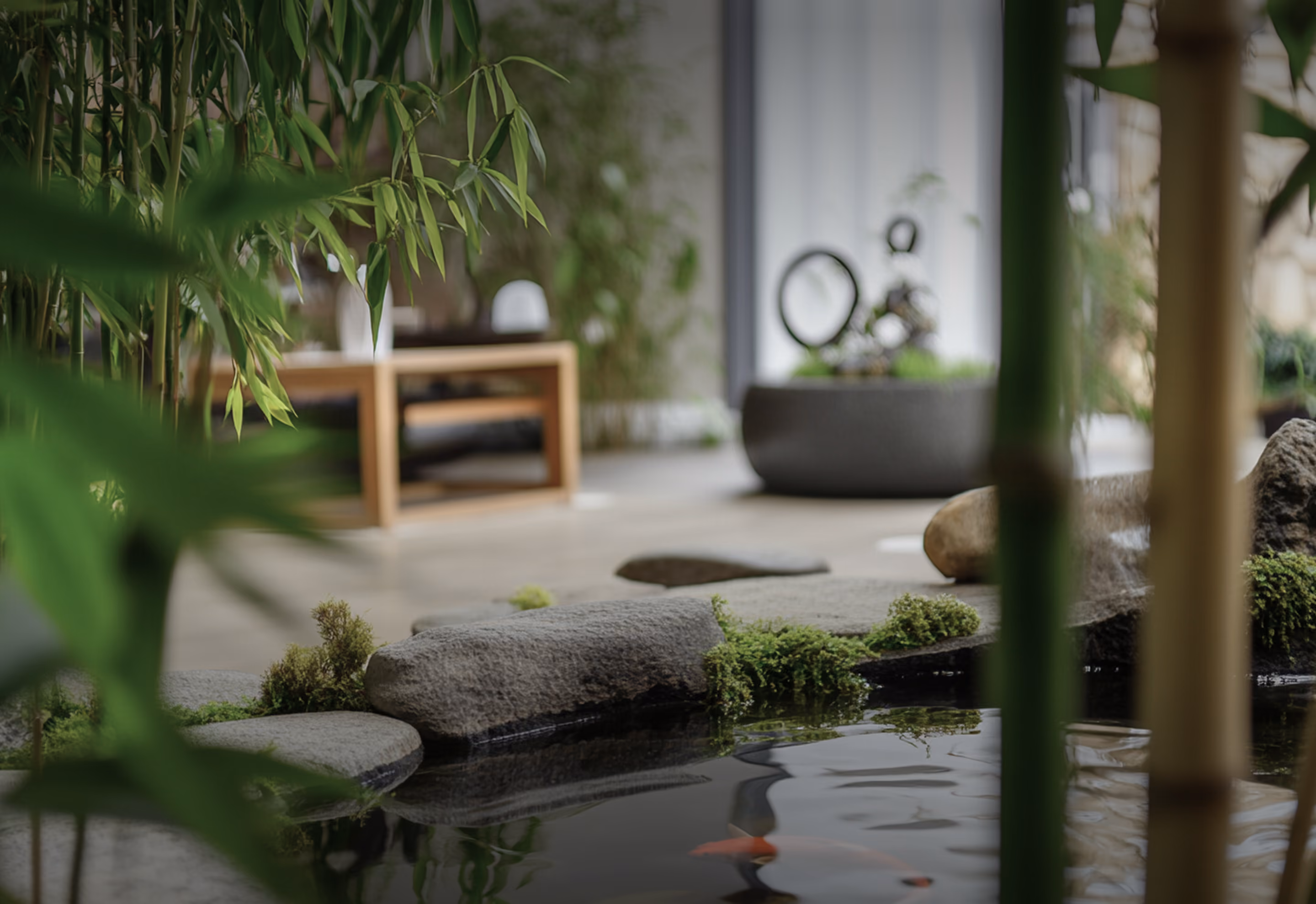 Peaceful indoor Zen garden with a small pond edged by smooth stones and patches of green moss, leafy bamboo stalks, and a wooden bench blurred in the background.