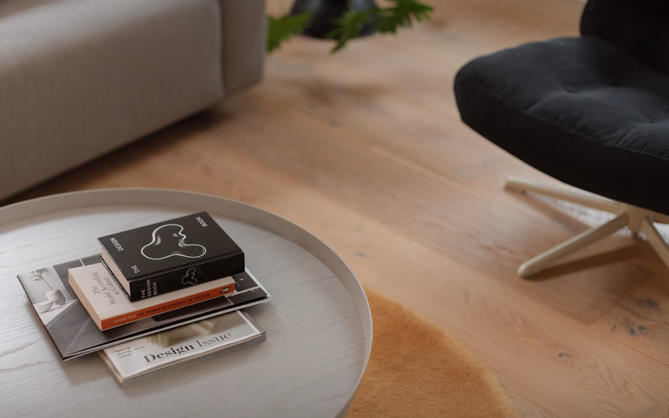 Round wooden coffee table with a stack of design and art books placed on top, positioned on a light wooden floor next to a beige sofa and a dark upholstered chair with white legs.