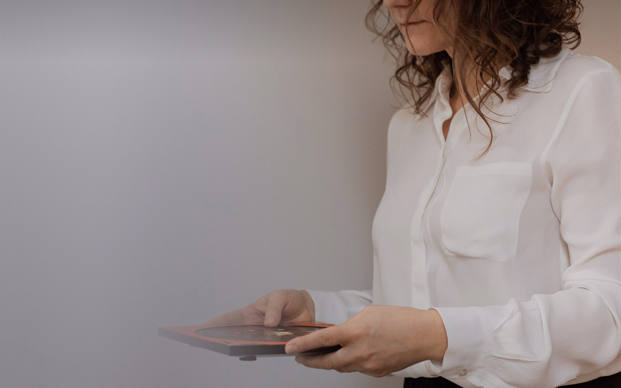 Woman in a white blouse holding and looking at a portable electronic device with a dark screen, against a plain background.