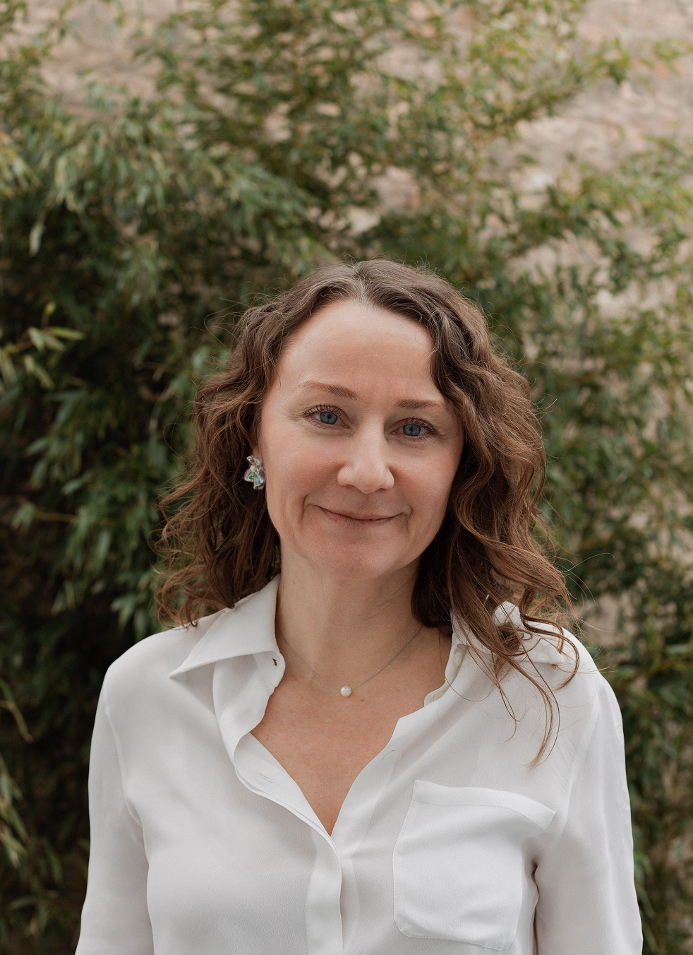 Portrait of a woman with wavy brown hair wearing a white button-up shirt and a small pendant necklace, standing outdoors with green foliage in the background.