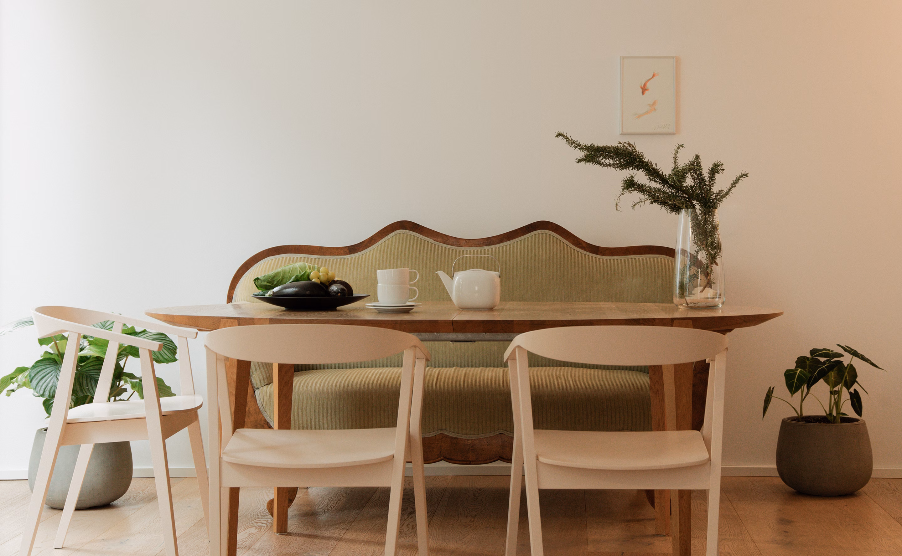 Minimalist dining area with a wooden table, two beige chairs, a vintage green cushioned bench, a teapot with a cup, a black tray with vegetables on the table, and green potted plants on either side.