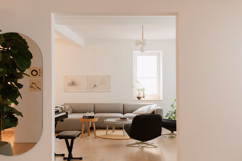 Minimalist living room with a gray sectional sofa against a white wall, a modern black armchair, two round coffee tables, and light wooden flooring, viewed through a rectangular doorway with a large green plant and a vertical mirror on the left wall.