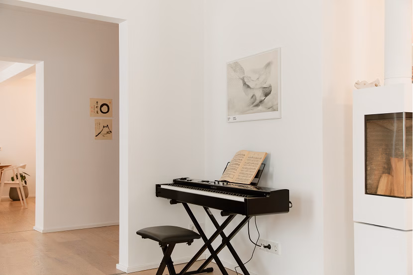 Small black digital piano on a folding stand with a matching black padded stool and sheet music placed on the piano, set against a white wall with a soft-toned drawing above it, next to a modern white fireplace in a bright room with light wood flooring.