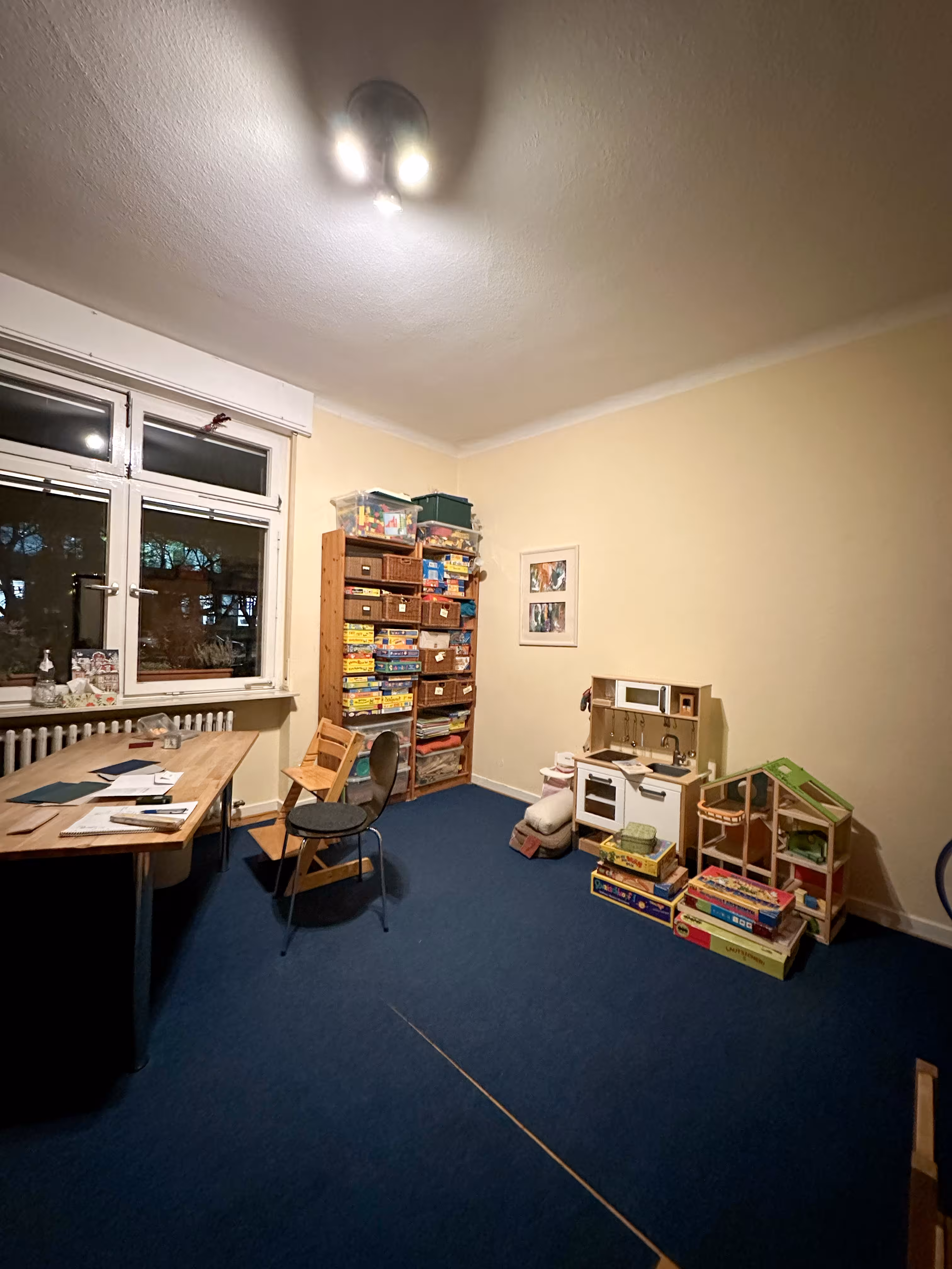 Room with blue carpet and cream walls containing a wooden table with papers and writing tools, two chairs, a tall shelf stacked with board games and storage baskets, a play kitchen set against the wall, and assorted toys and board games on the floor.