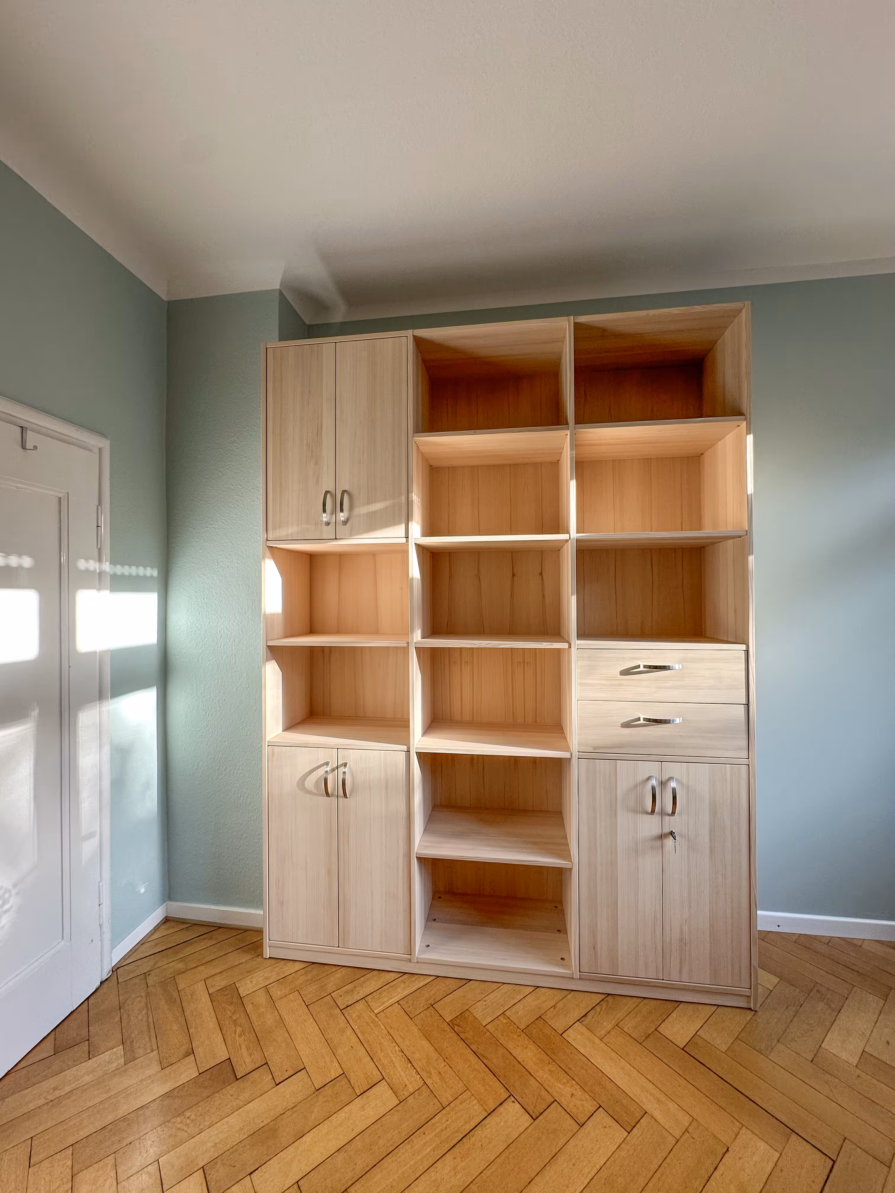 Tall wooden shelving unit with multiple open square compartments, two drawers, and cabinets with silver handles, standing against a light blue wall on herringbone wood flooring near a white door with sunlight casting shadows.