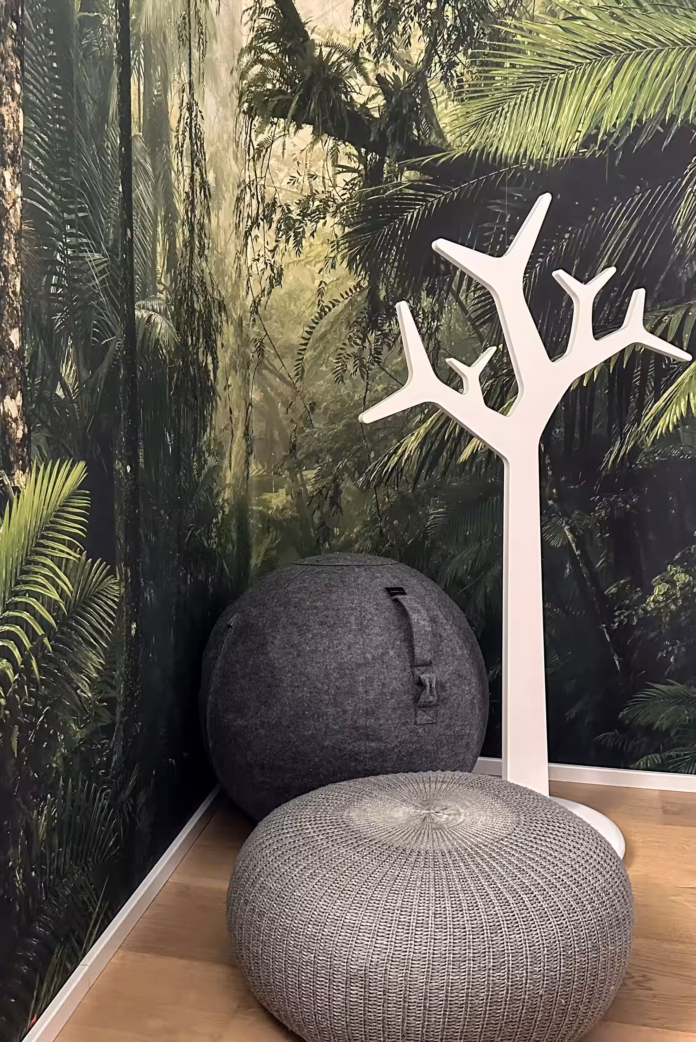 Corner of a room with a nature-themed wall mural featuring dense green foliage, a round dark gray felt pouf, a round light gray knitted ottoman, and a white modern coat rack shaped like tree branches.