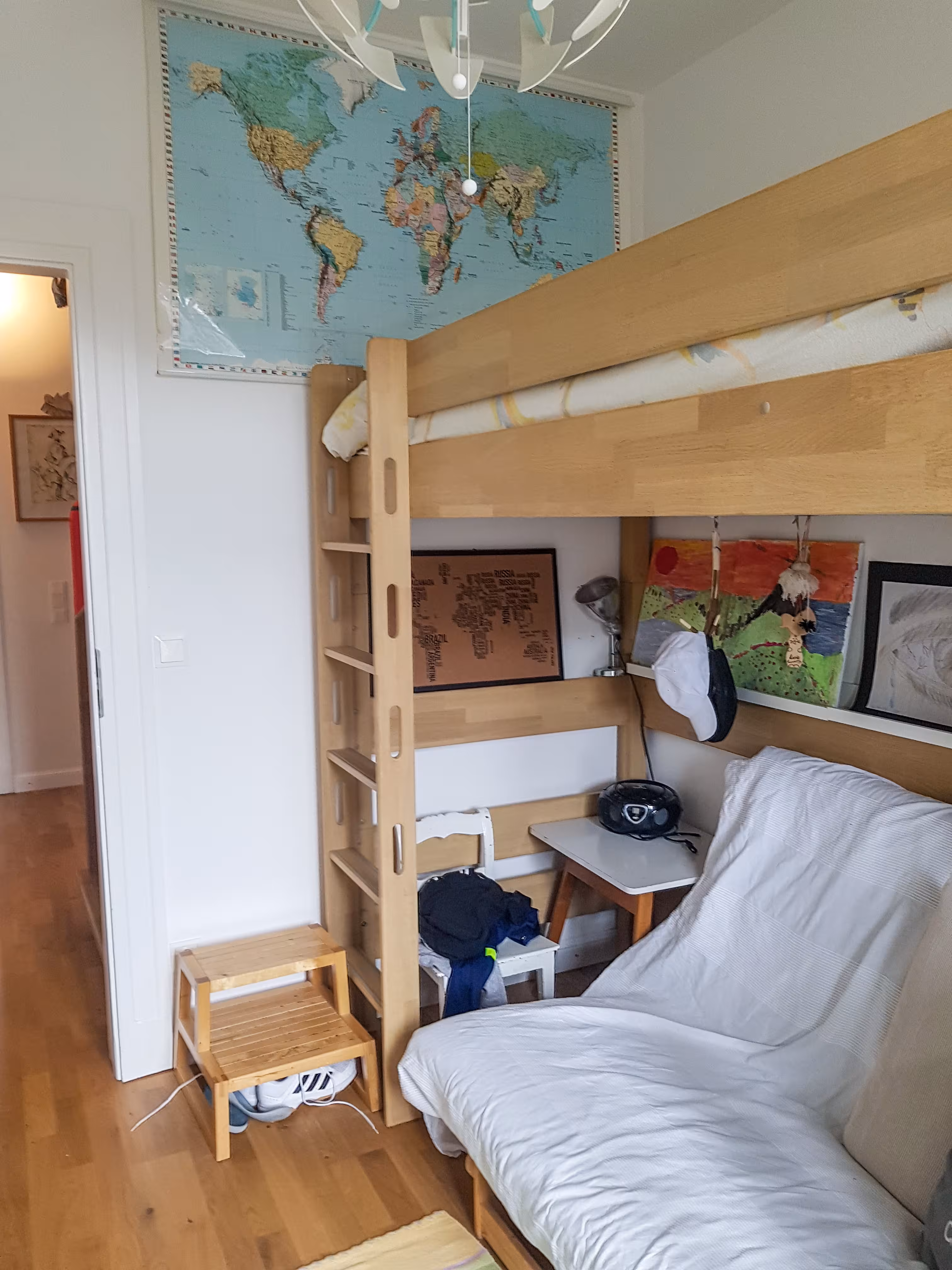 A small bedroom with a wooden loft bed, a futon sofa below, a small side table holding a black radio, and children's artwork hanging on the wall. A world map poster is mounted high on the wall above the bed. Wooden floor and a small step stool with shoes underneath are visible near the doorway.