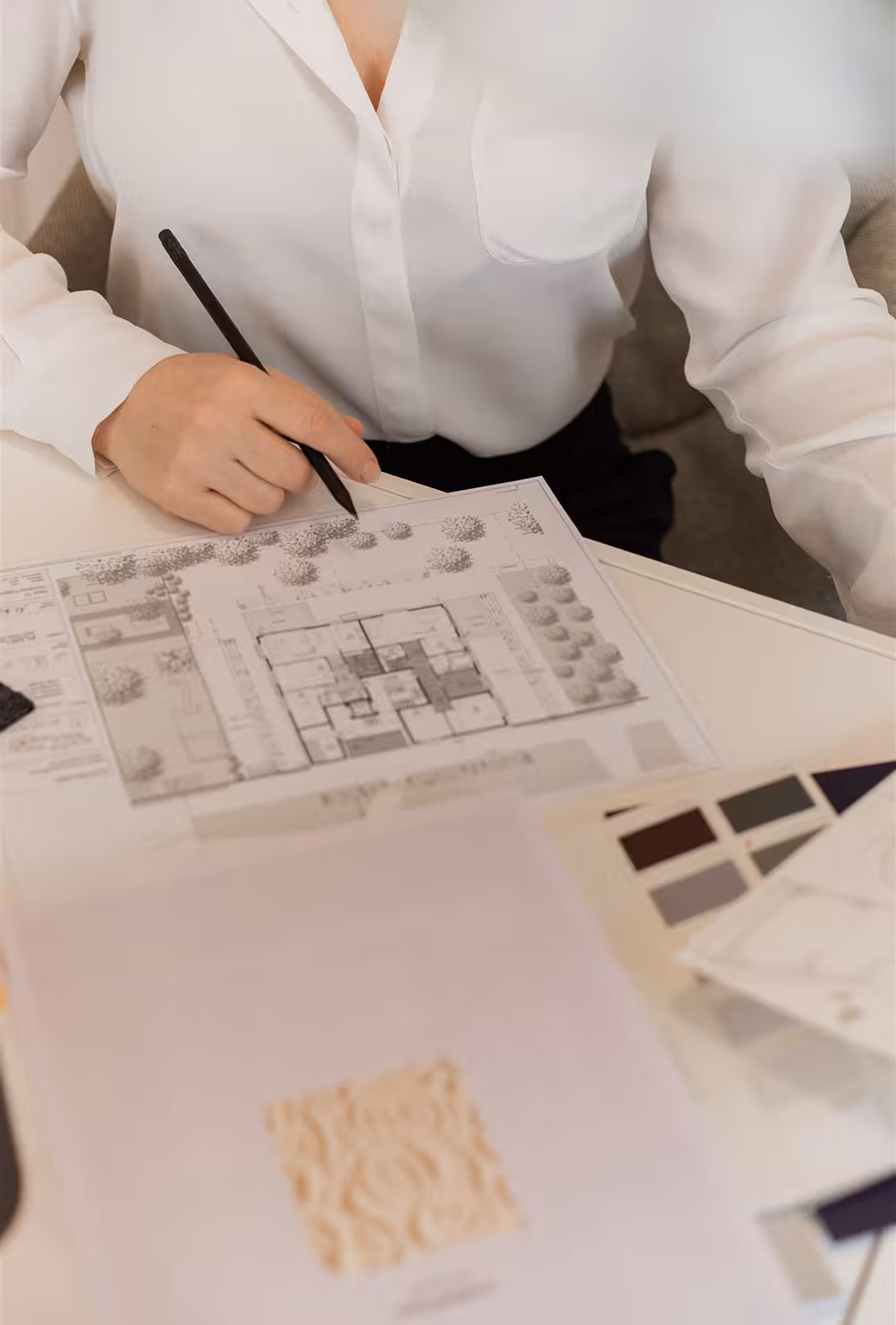Person in a white shirt holding a black pencil and pointing at an architectural floor plan on a desk with color swatches and textured material samples.