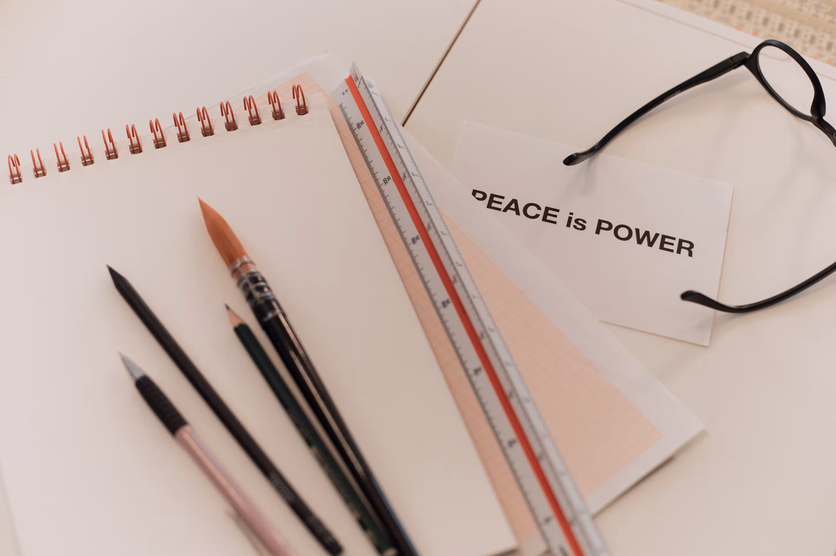 A white desk with a blank spiral notebook, several pencils and a paintbrush, a ruler, a pair of black-rimmed glasses, and a card with the text 'PEACE is POWER'.