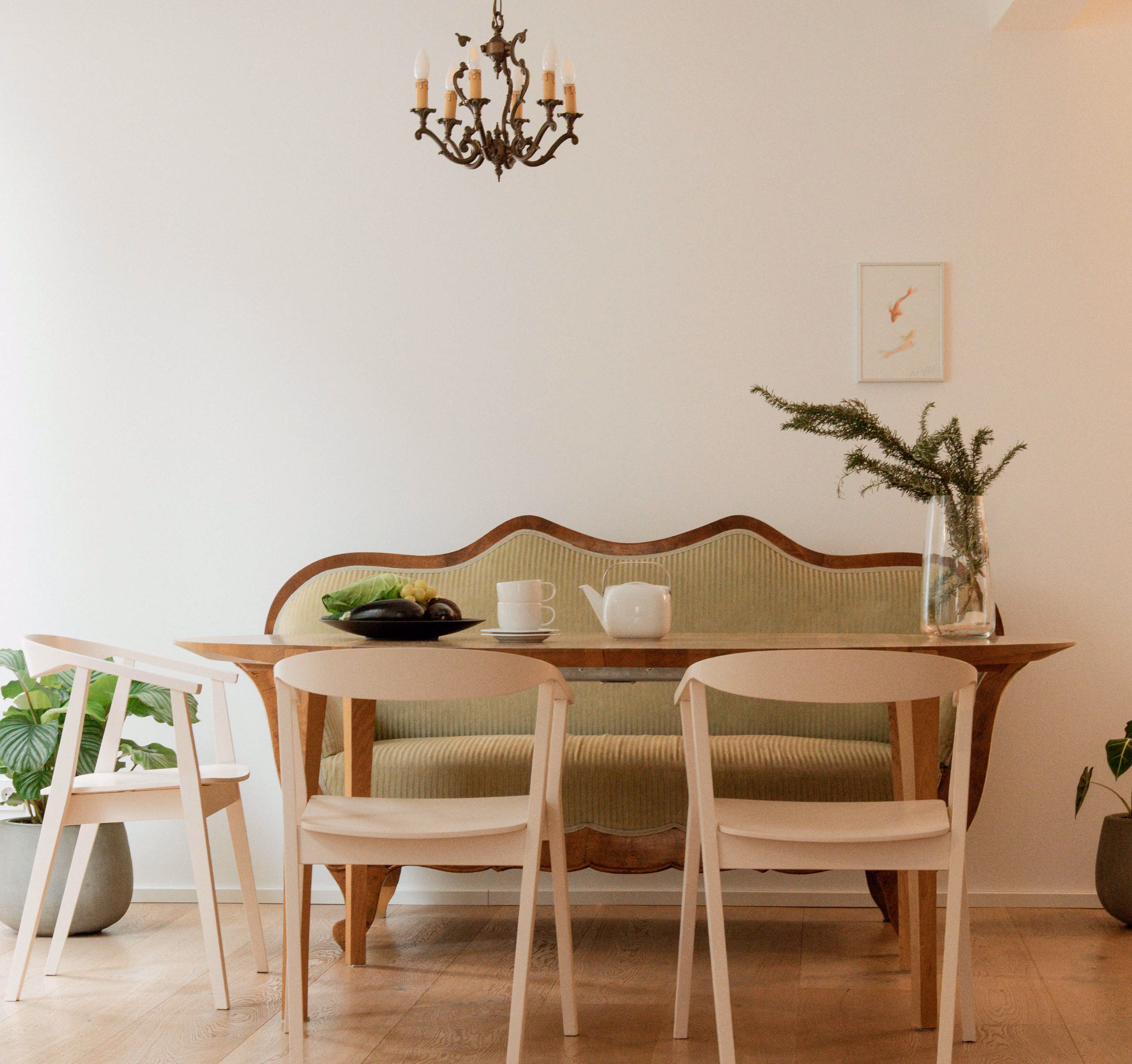 Dining area with a wooden table set in front of a vintage-style wooden bench with green upholstery, accompanied by three modern minimalist light wood chairs, a chandelier above, and plants on either side.