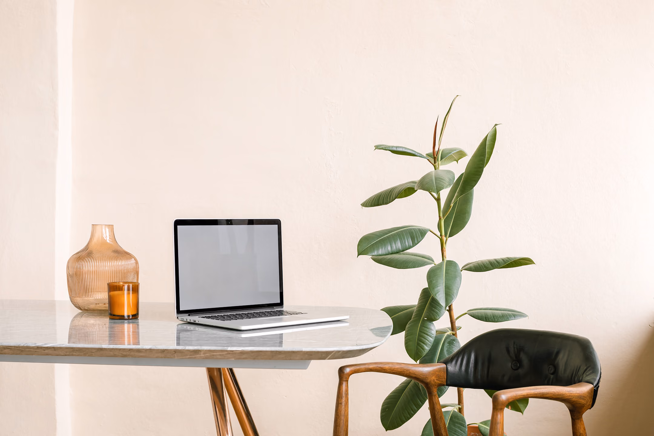 Minimalistic workspace with a silver laptop on a round marble table, a brown glass vase and candle to the left, and a green leafy plant next to a black chair with wooden armrests against a beige wall.