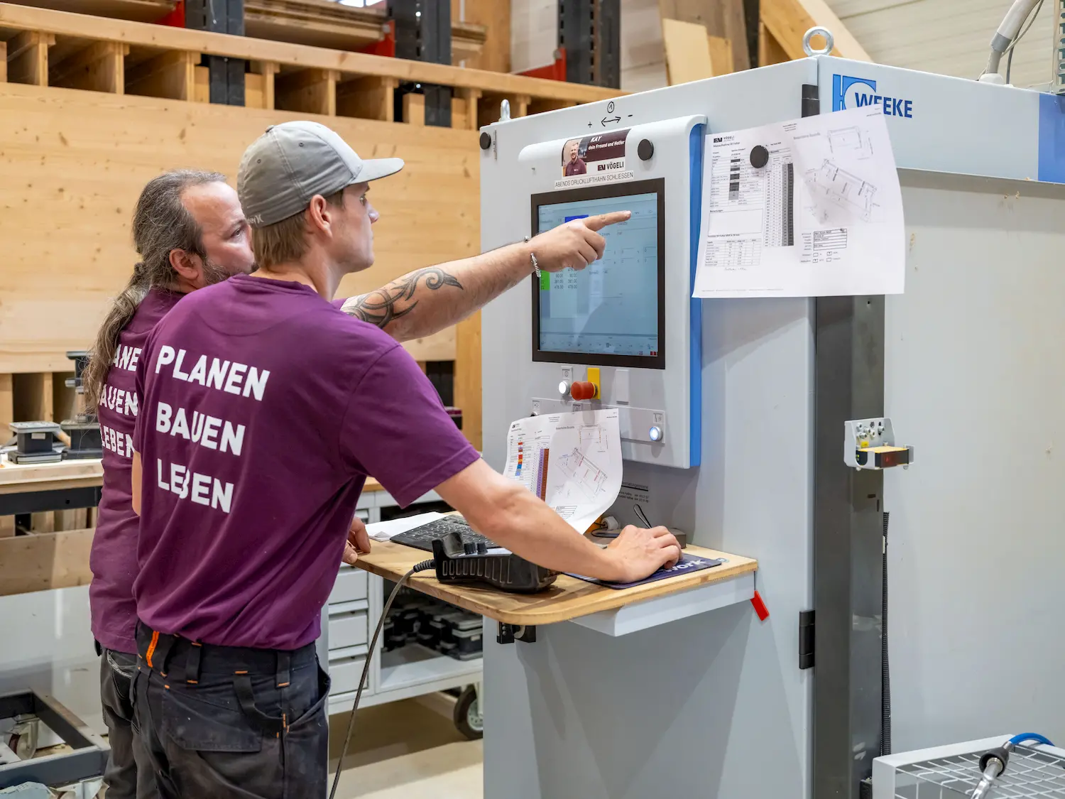 Zwei Männer bedienen gemeinsam eine CNC-Maschine und betrachten technische Pläne auf einem Bildschirm.