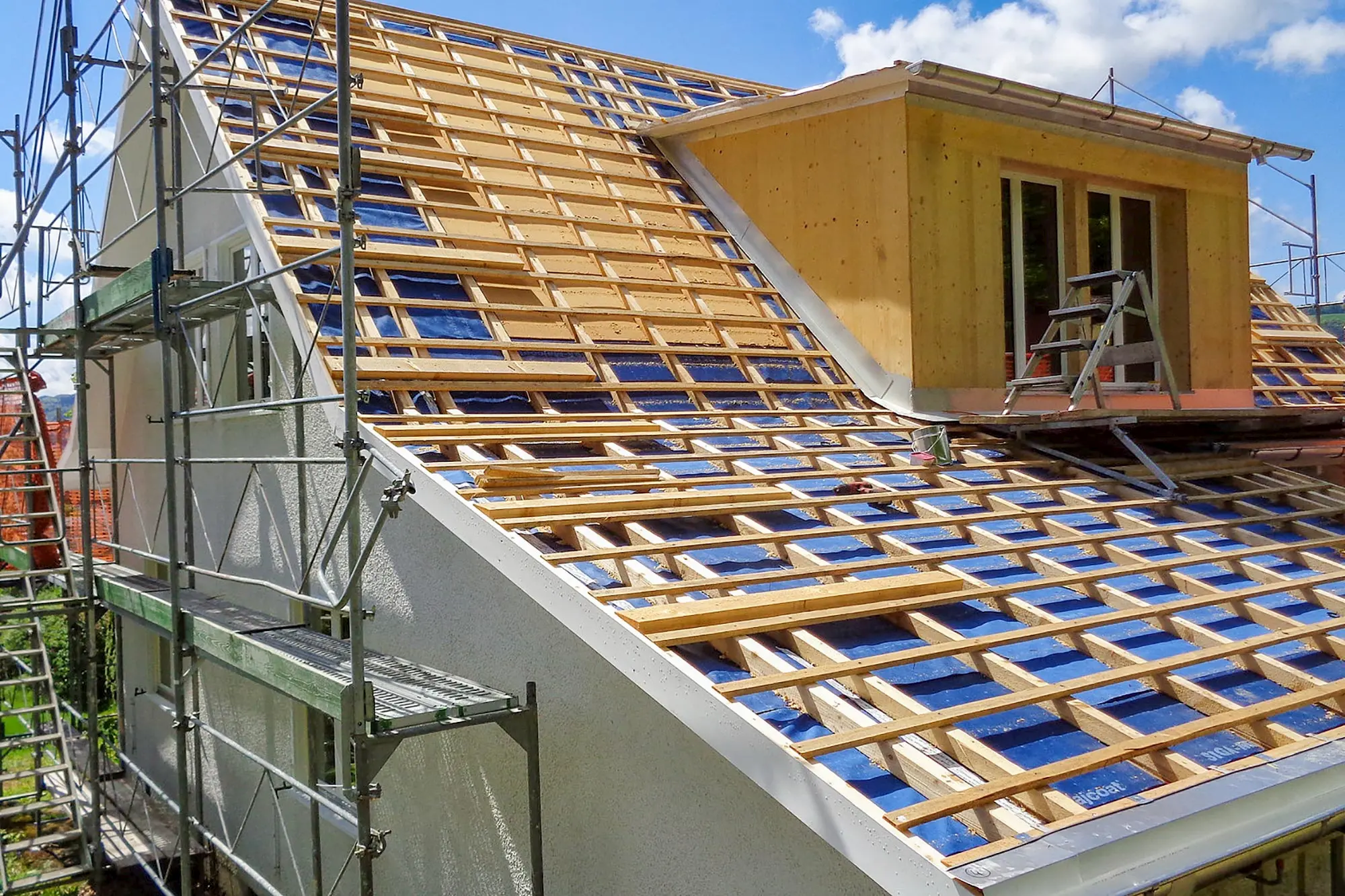 Hausdach während einer Sanierung, mit Gerüst und sichtbarer Holzlattung, über die neue Dacheindeckung verlegt wird; im oberen Bereich ein Dachaufbau mit Fenster.
