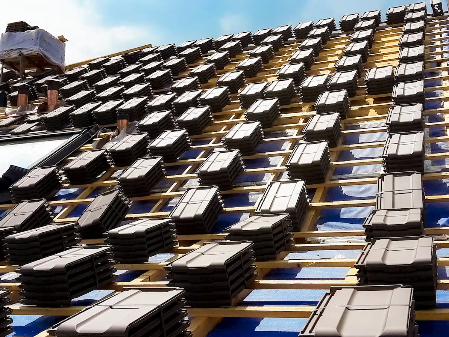 Gestapelte schwarze Dachziegel liegen in Reihen auf einer vorbereiteten Dachlattung mit blauer Unterspannbahn.