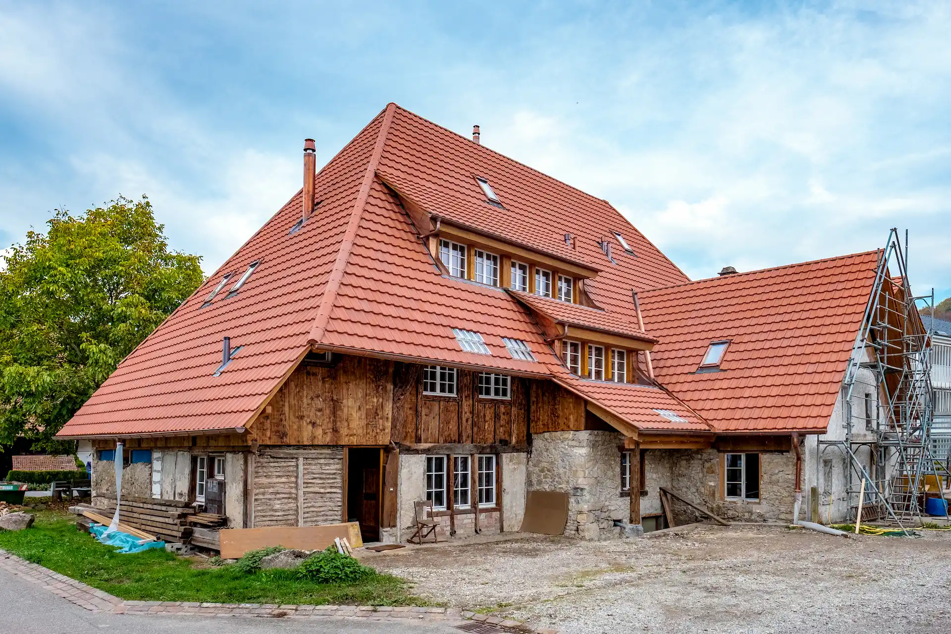 Historisches Bauernhaus mit neu gedecktem roten Ziegeldach, teils aus Holz, teils aus Naturstein gebaut, vor einem blauen Himmel. Vor dem Haus liegt Baumaterial, rechts ist ein Gerüst angebracht.