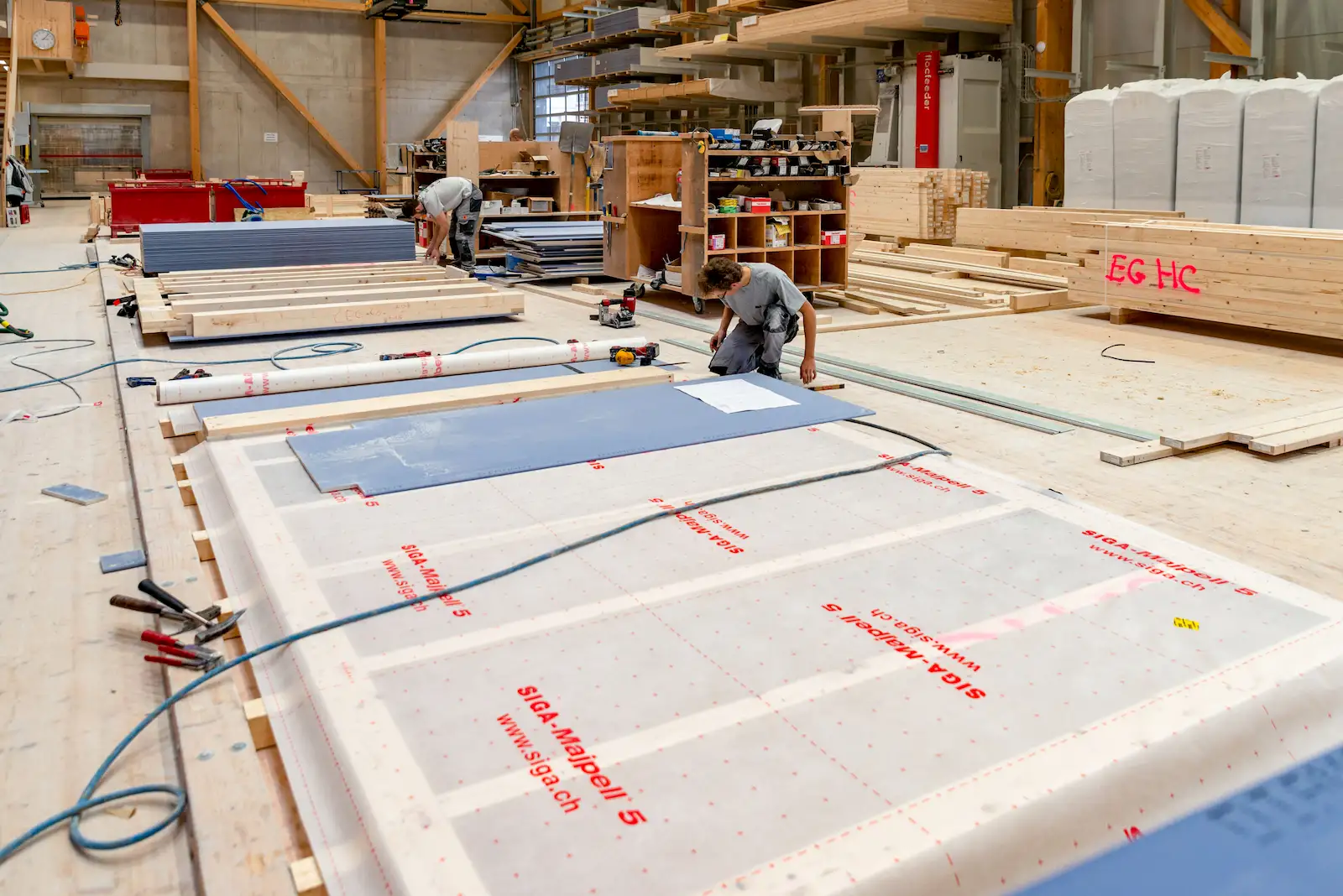 Zwei Handwerker in einer Werkhalle fertigen Holzelemente für den Hausbau vor. Auf dem Boden liegen Holzrahmen, Dämmmaterial und Baupläne, umgeben von Werkzeugen und Materialien.