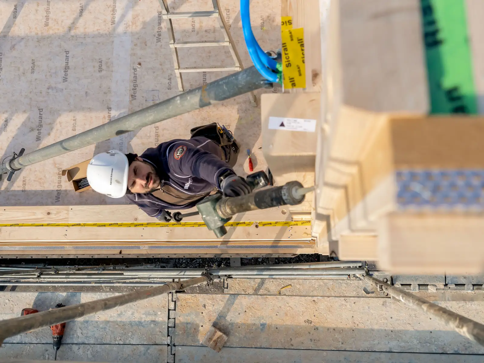Bauarbeiter mit Schutzhelm befestigt von einem Gerüst aus Bauteile eines Holzhauses. Im Vordergrund sind Holzelemente und Bauwerkzeuge sichtbar.