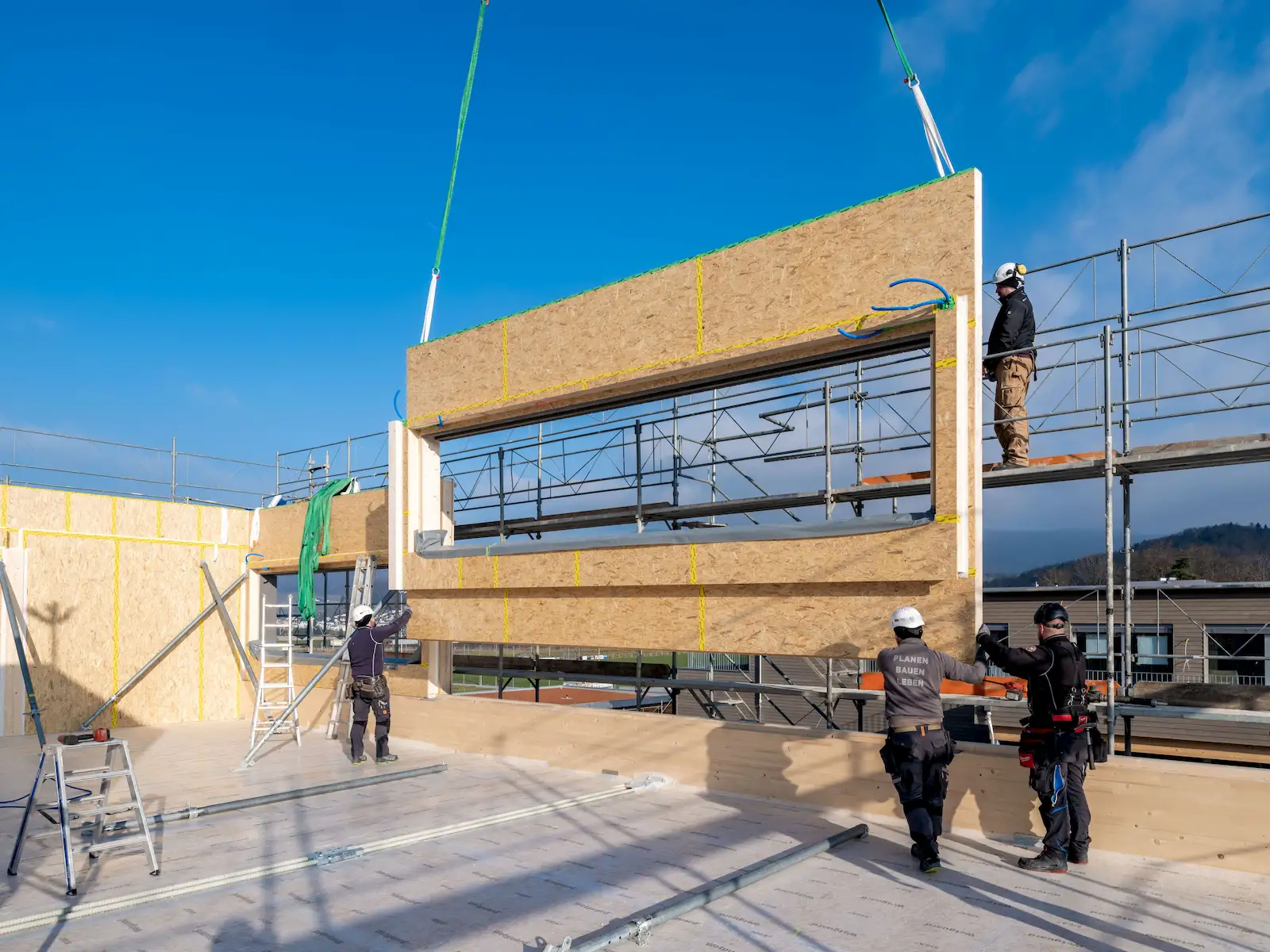 Arbeiter montieren mit einem Kran eine große vorgefertigte Holzwand mit Fensteröffnung auf einer Baustelle. Mehrere Personen in Schutzkleidung und Helmen sichern das Element.