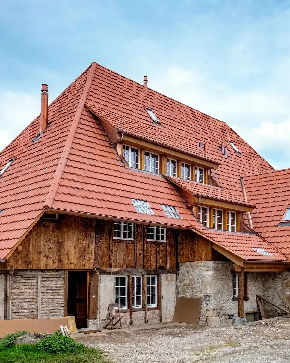 Historisches Bauernhaus mit neu gedecktem roten Ziegeldach, teils aus Holz, teils aus Naturstein gebaut, vor einem blauen Himmel. Vor dem Haus liegt Baumaterial, rechts ist ein Gerüst angebracht.