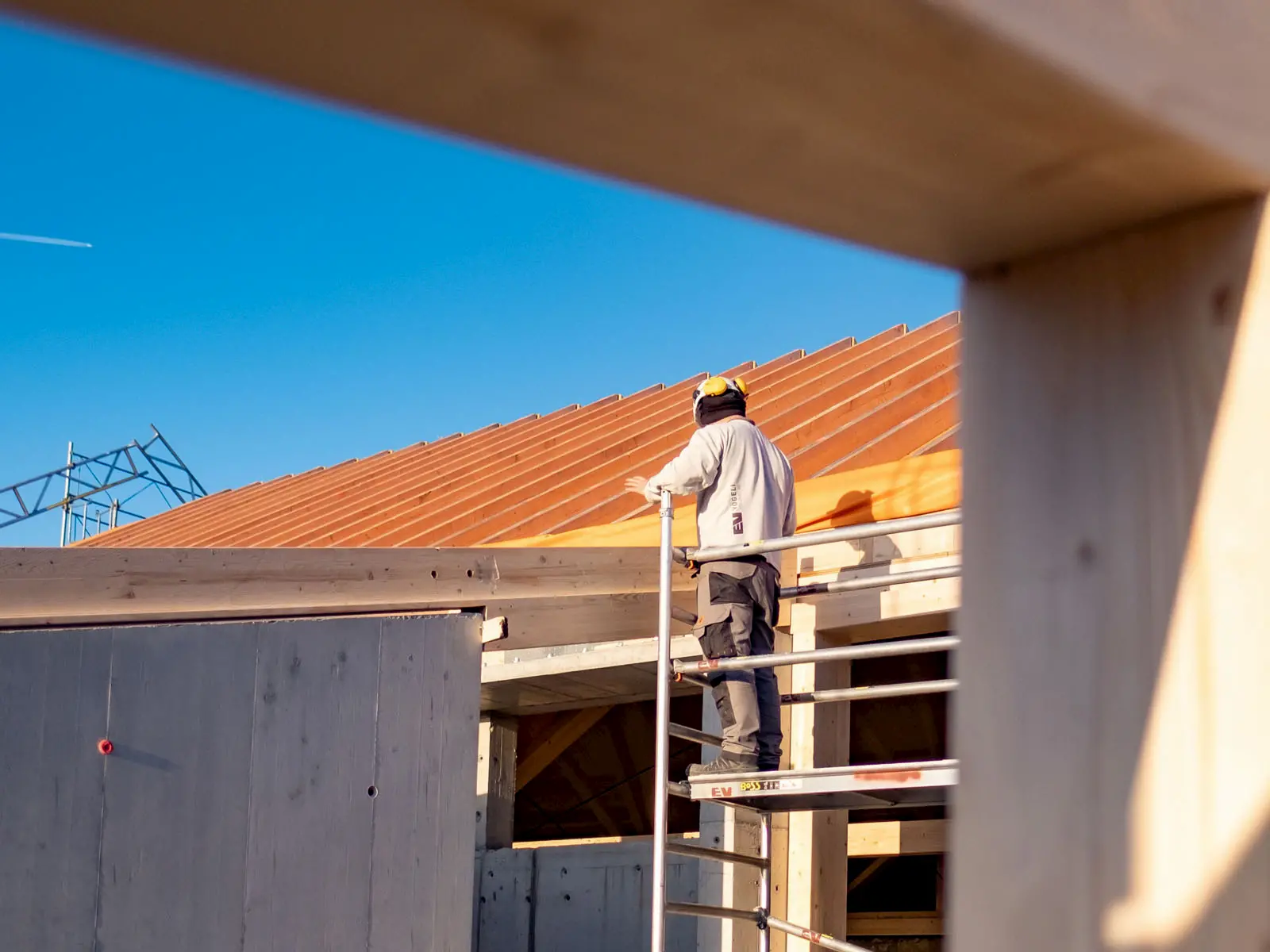 Zimmermann auf Gerüst montiert Holzdach unter blauem Himmel, sorgfältige Ausführung nachhaltiger Holzkonstruktion.