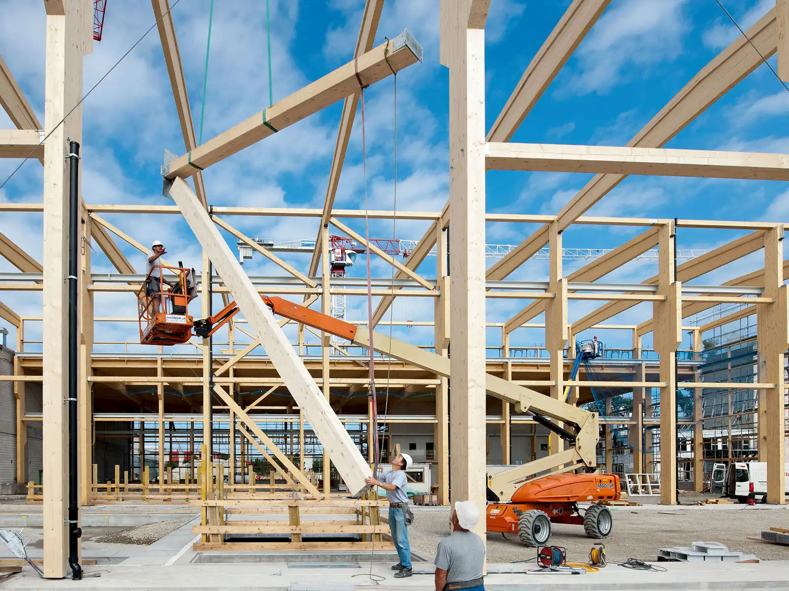 Montage von Holztragwerk auf Baustelle, Kräne und Arbeitsbühnen im Einsatz – effiziente Konstruktion moderner Hallenstruktur.