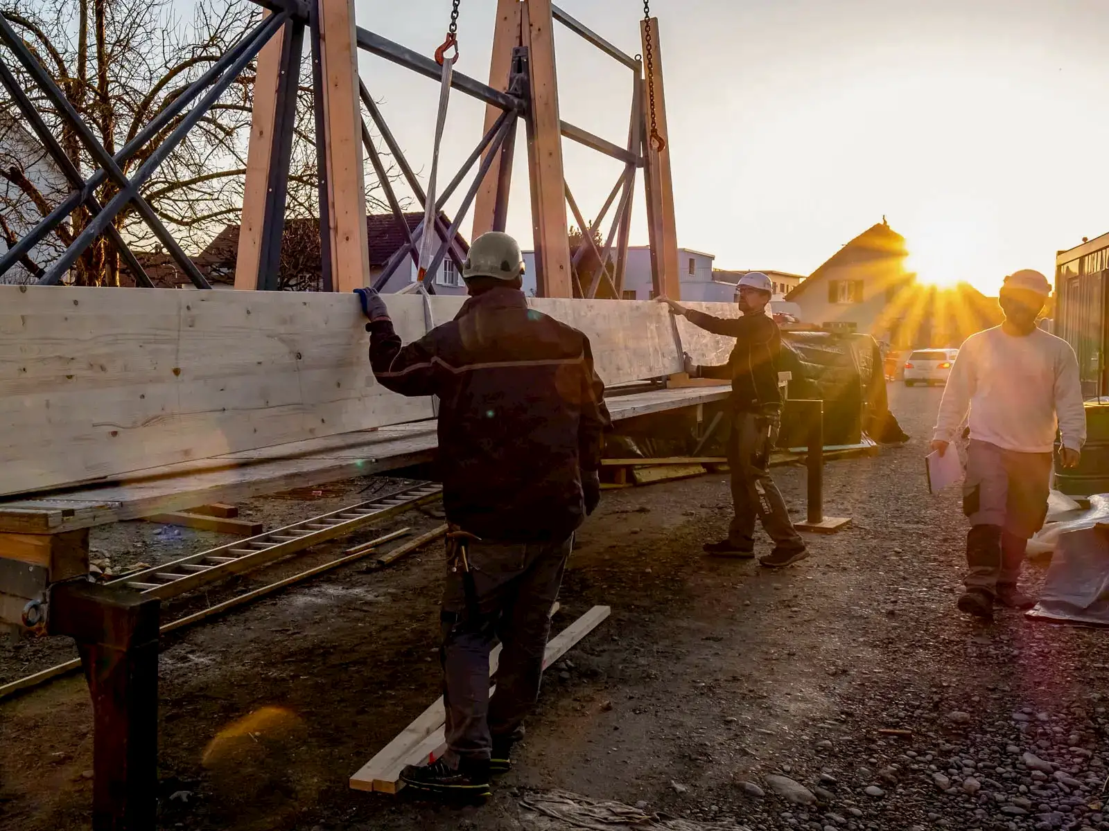 Team hebt großen Holzträger bei Sonnenuntergang, präzise Montagearbeit auf Baustelle mit stimmungsvoller Lichtkulisse.