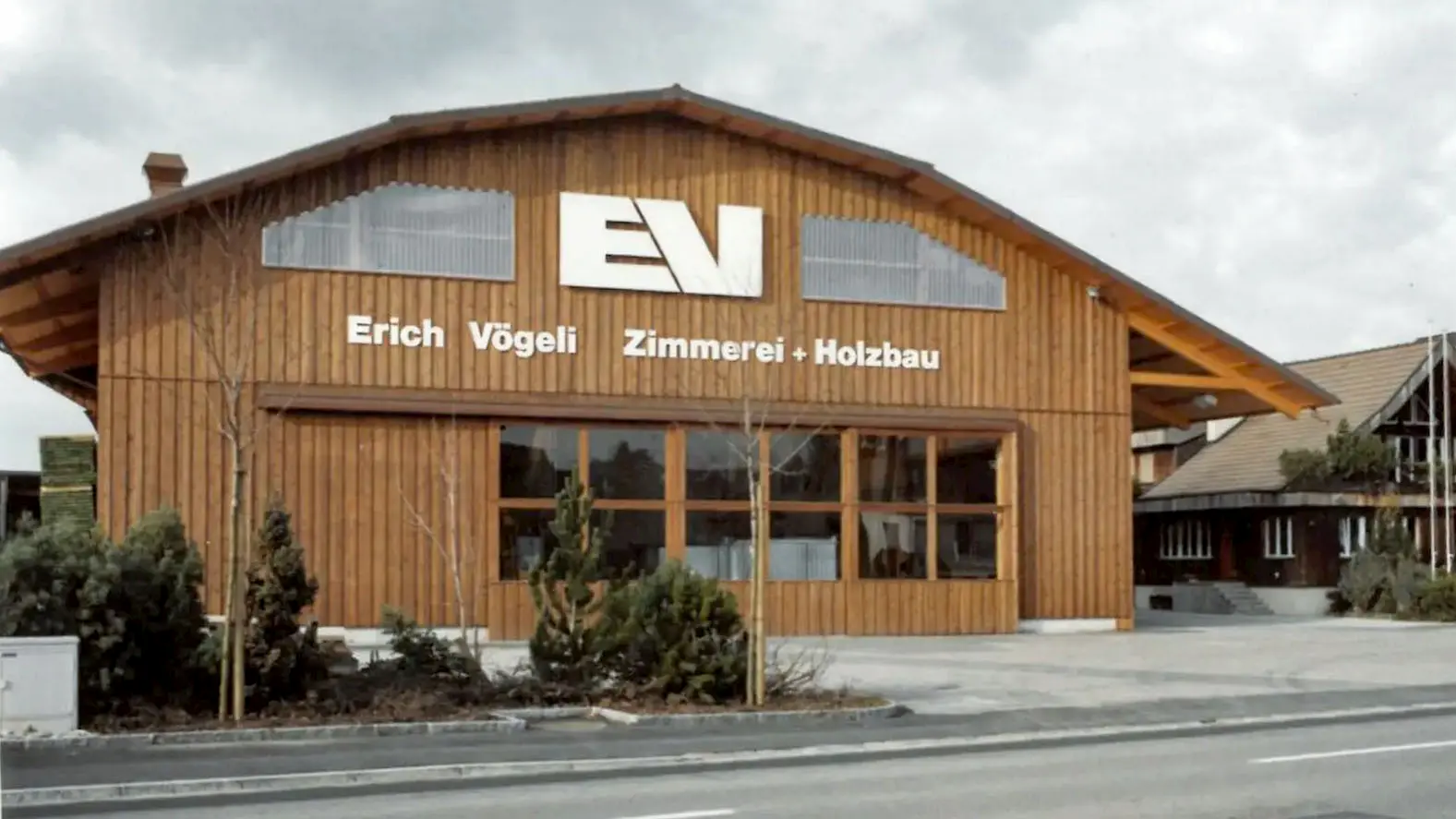 Holzgebäude mit Schild 'Erich Vögeli Zimmerei + Holzbau' und großem Firmenlogo 'EV' an der Fassade. Fertiger Erweiterungsbau 1986