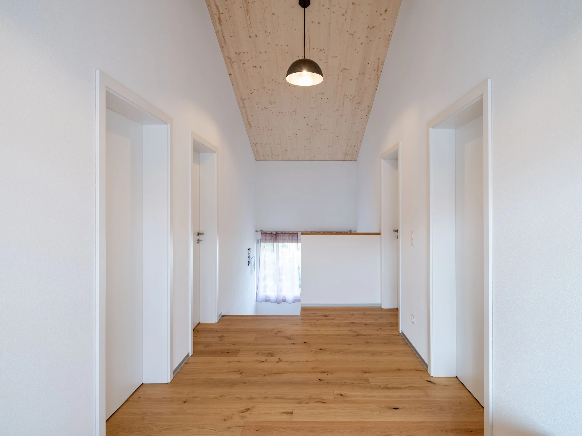 Heller, moderner Hausflur mit weiß gestrichenen Wänden, vier weißen Türen und einem Boden aus geölten Eichedielen mit dunklen Astfüllungen. Die Decke besteht aus hellem Holz mit sichtbarer Maserung und einer zentral hängenden Lampe. Am Ende des Flurs befindet sich ein kleines Fenster mit transparentem Vorhang