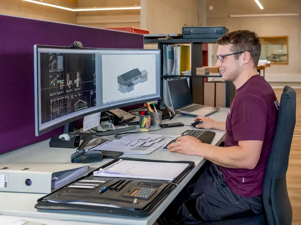 Ein Mann sitzt am Schreibtisch und arbeitet am Computer mit CAD-Software, auf dem Bildschirm ist ein 3D-Hausmodell zu sehen.