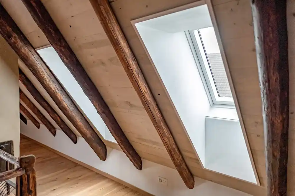 Dachschräge mit sichtbaren Holzbalken und zwei Dachfenstern, die Tageslicht hereinlassen.