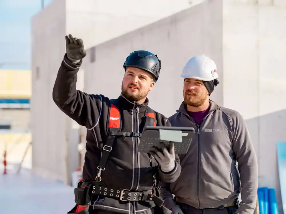 Zwei Bauarbeiter mit Schutzhelmen und Tablet besprechen sich auf einer Baustelle.