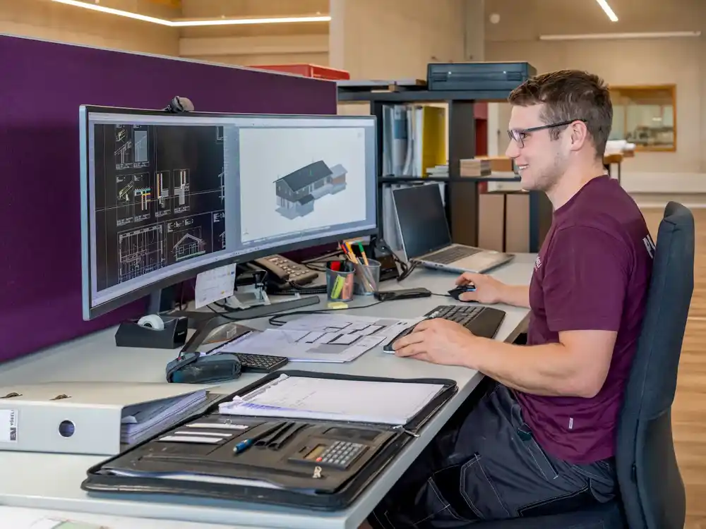 Mann arbeitet an Computermonitor mit Hausplänen und Architekturzeichnungen im Büro.
