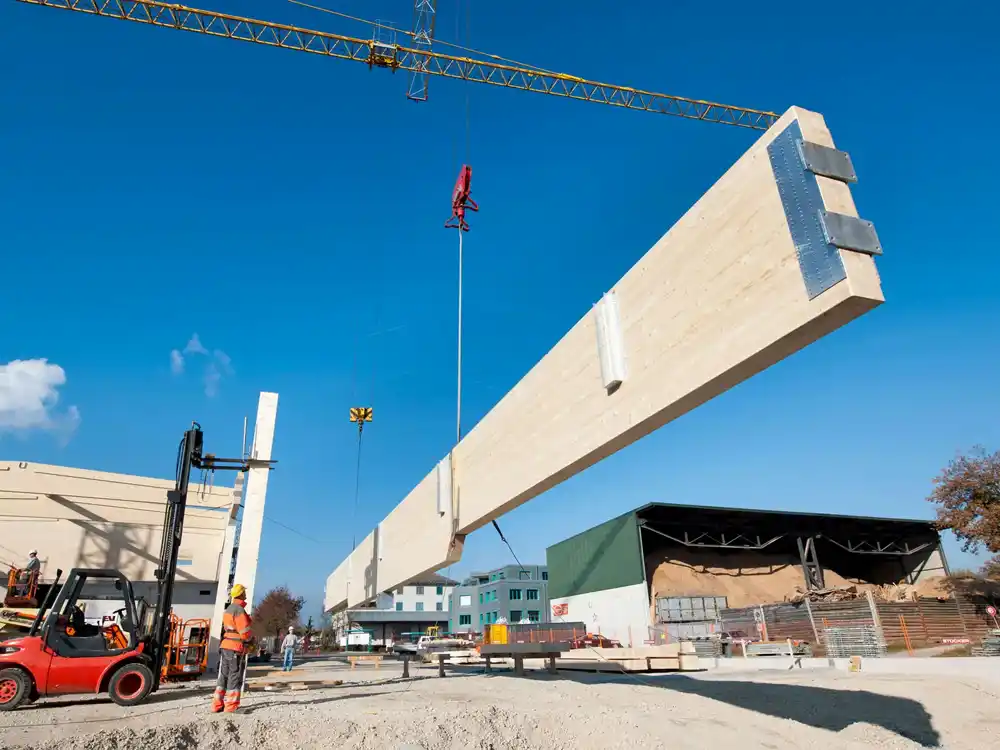 Baustelle mit einem großen Holzträger, der von einem Kran gehoben wird, und Bauarbeitern bei der Arbeit.