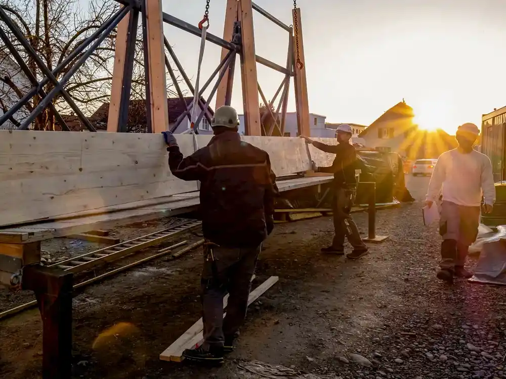 Drei Bauarbeiter mit Helmen heben ein grosses Holzbrett an einer Baustelle bei Sonnenuntergang.