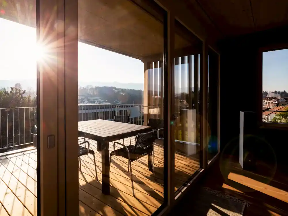 Sonnendurchflutete Terrasse mit Holzrostboden, Tisch und vier Stühlen, Blick auf Landschaft und Häuser.