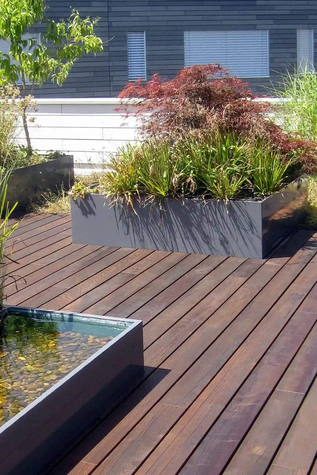 Moderne Holzboden-Terrasse mit rechteckigen Pflanzkästen und Wasserbecken, umgeben von Pflanzen.