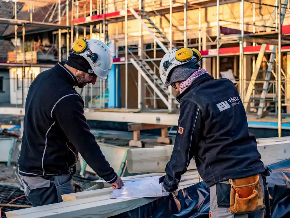 Zwei Bauarbeiter mit Schutzhelmen überprüfen Baupläne auf einer Baustelle mit Gerüst im Hintergrund.