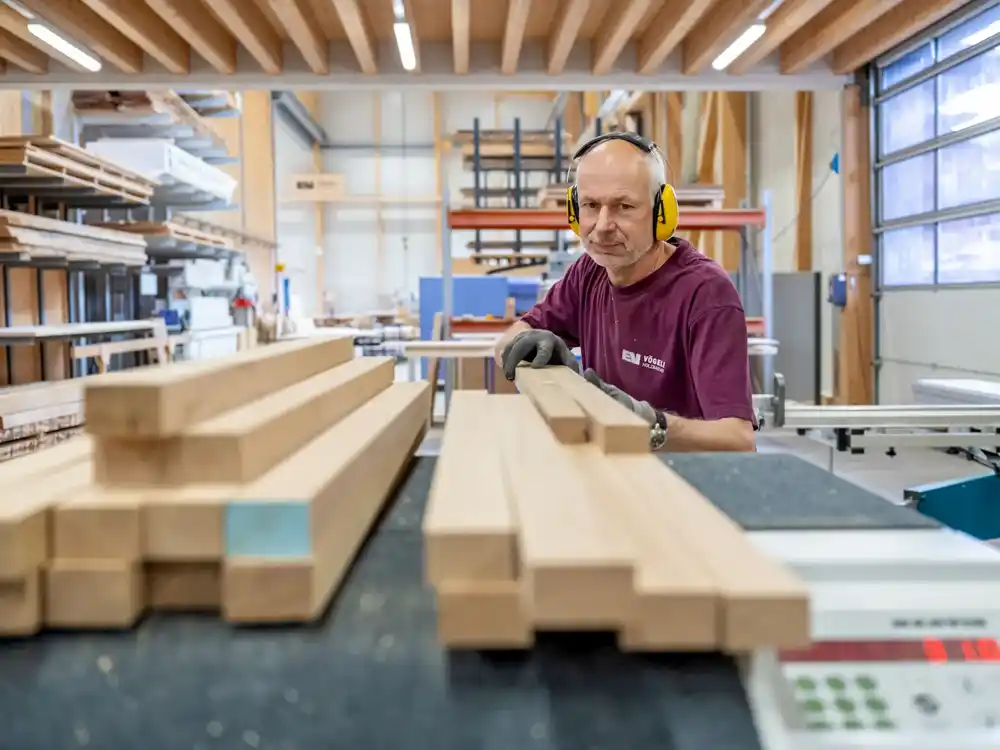 Ein Tischler mit Gehörschutz arbeitet in einer Holzwerkstatt mit mehreren Holzlatten.