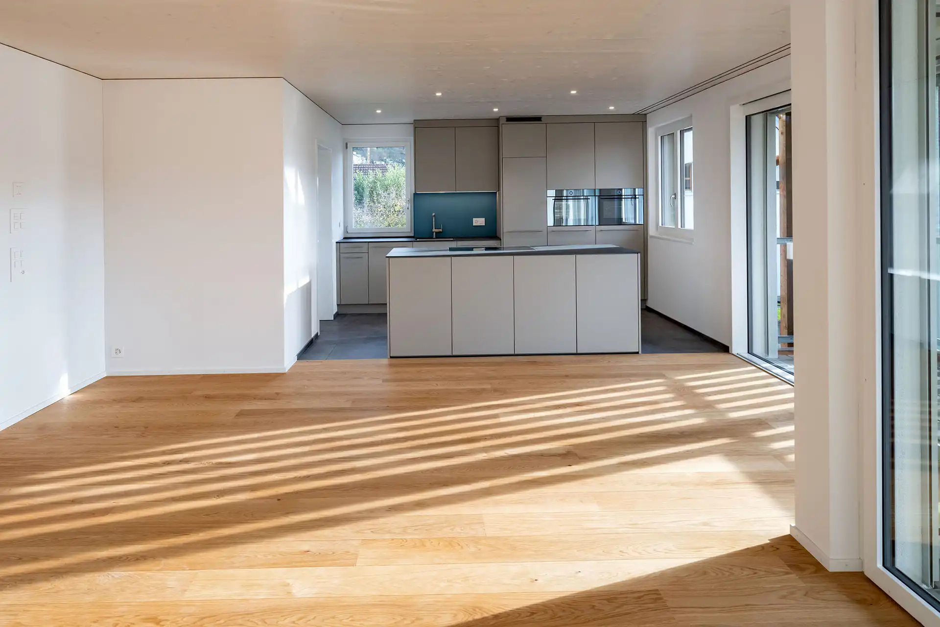 Helle Wohnküche mit Holzparkettboden, großen Fenstern und moderner grauer Küchenzeile mit Insel in einem Neubau.