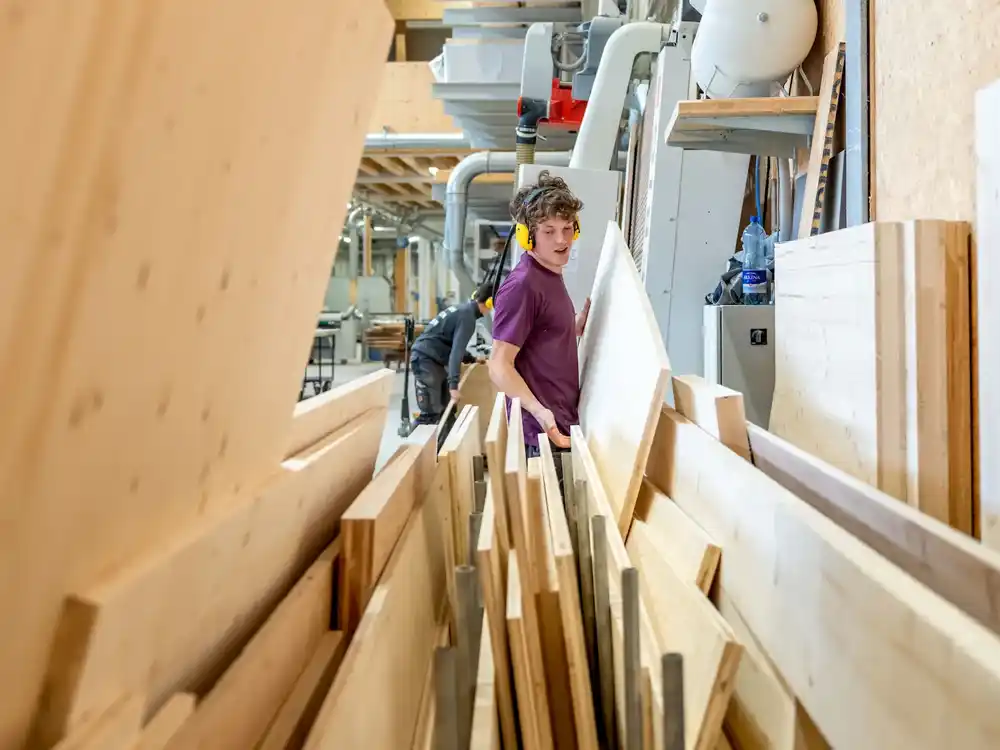 Holzarbeiter trägt ein großes Holzbrett in einer Holzwerkstatt mit mehreren Holzplatten.