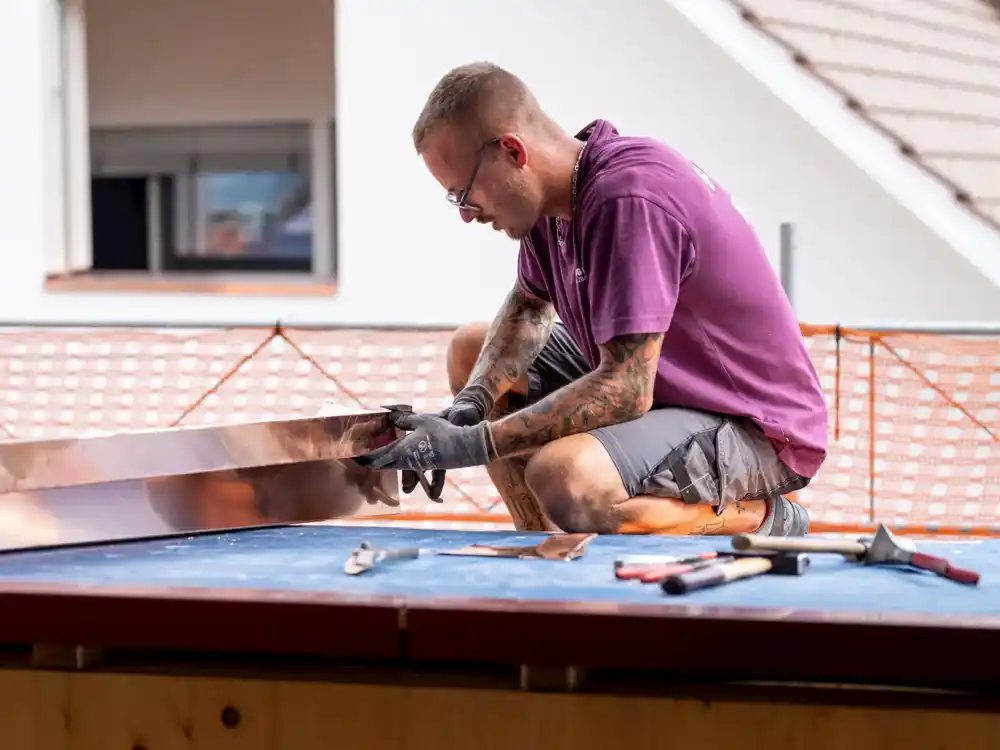 Handwerker mit Tattoos und Handschuhen bearbeitet ein Metallblech auf einer Arbeitsfläche mit Werkzeugen.