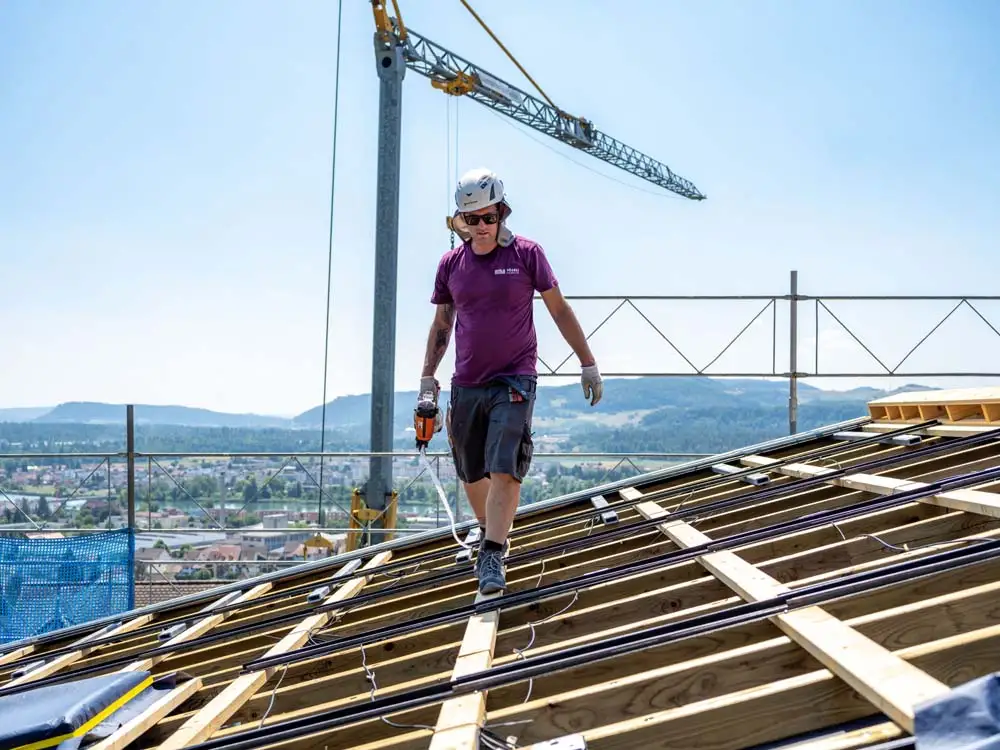 Bauarbeiter mit Helm und Bohrmaschine auf schrägem Holzgerüst bei sonnigem Himmel.