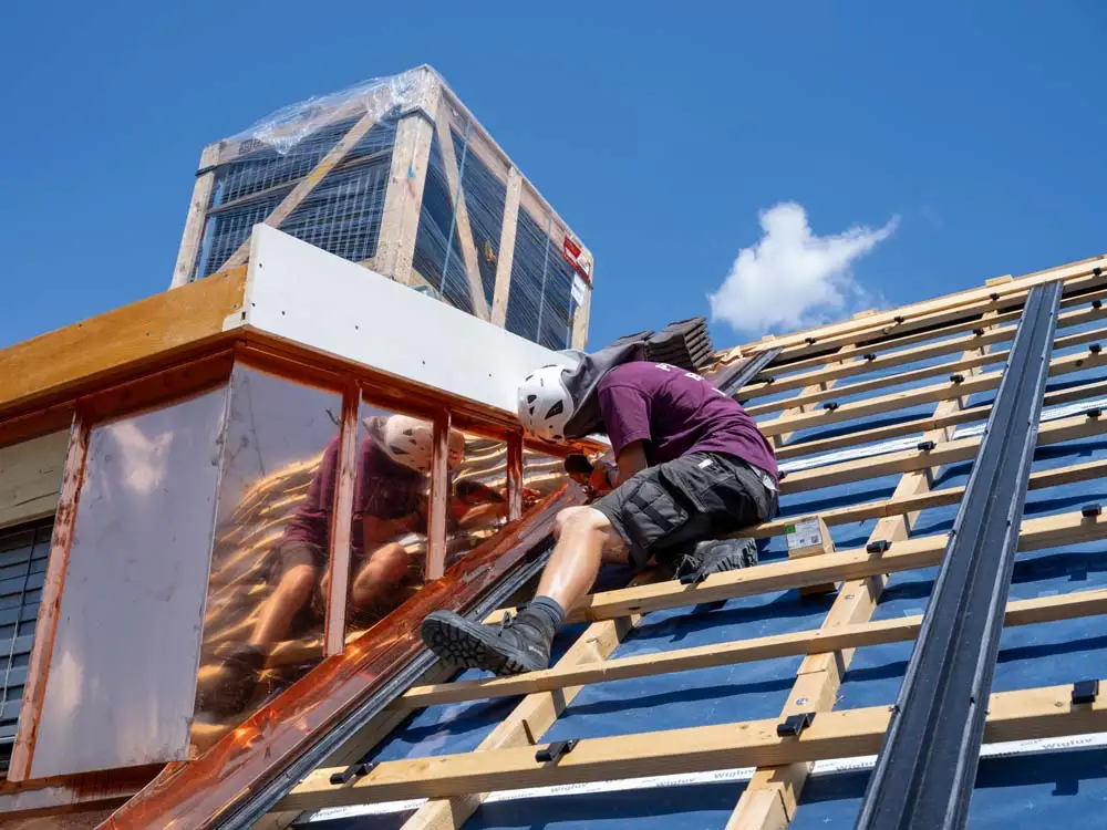 Dachdecker mit Helm arbeitet auf einem Holzgerüst an einem Dach unter blauem Himmel.