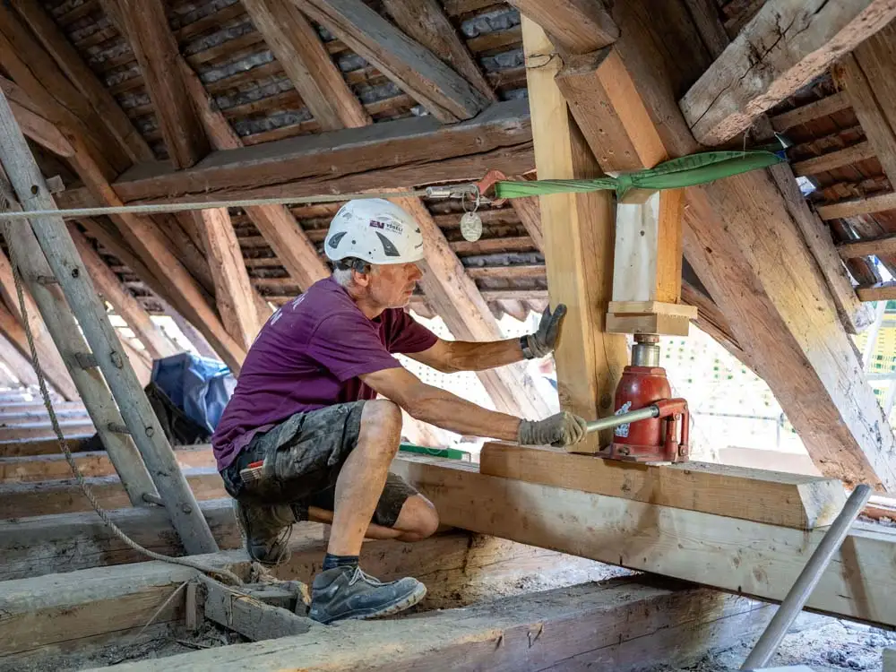 Handwerker mit Helm und Handschuhen arbeitet an Holzkonstruktion im Dachstuhl mit Hydraulikheber.