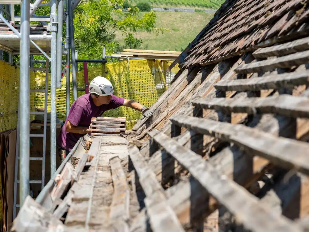 Handwerker mit Helm repariert ein Schindeldach mit Holzlatten und Ziegeln auf einem Gerüst.