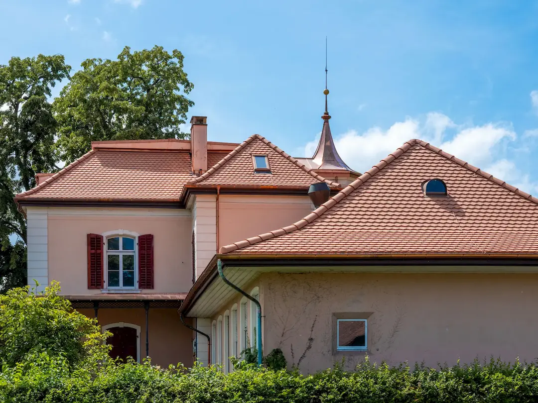 Haus mit roten Ziegeldächern, Dachfenstern und grünem Bewuchs davor bei blauem Himmel.