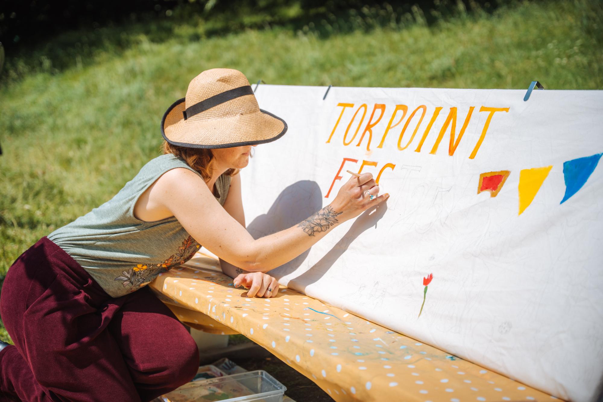 A lady paints a Torpoint Fest '25 sign
