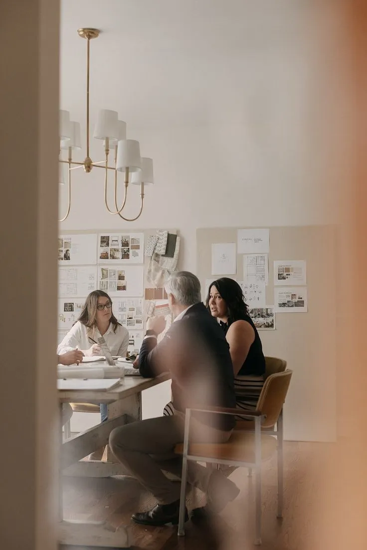 Three people sitting around a table in a meeting room with sketches and design plans on the wall.