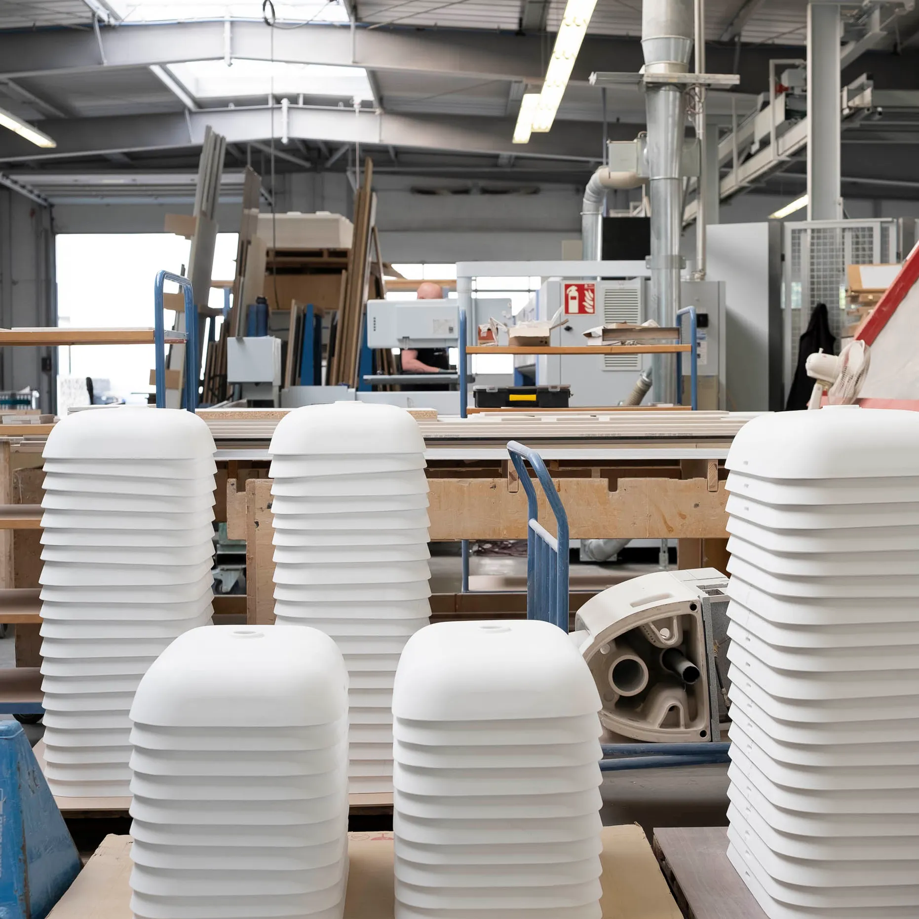 Stacks of white composite materials or parts in a workshop with machinery and wooden structures in the background.